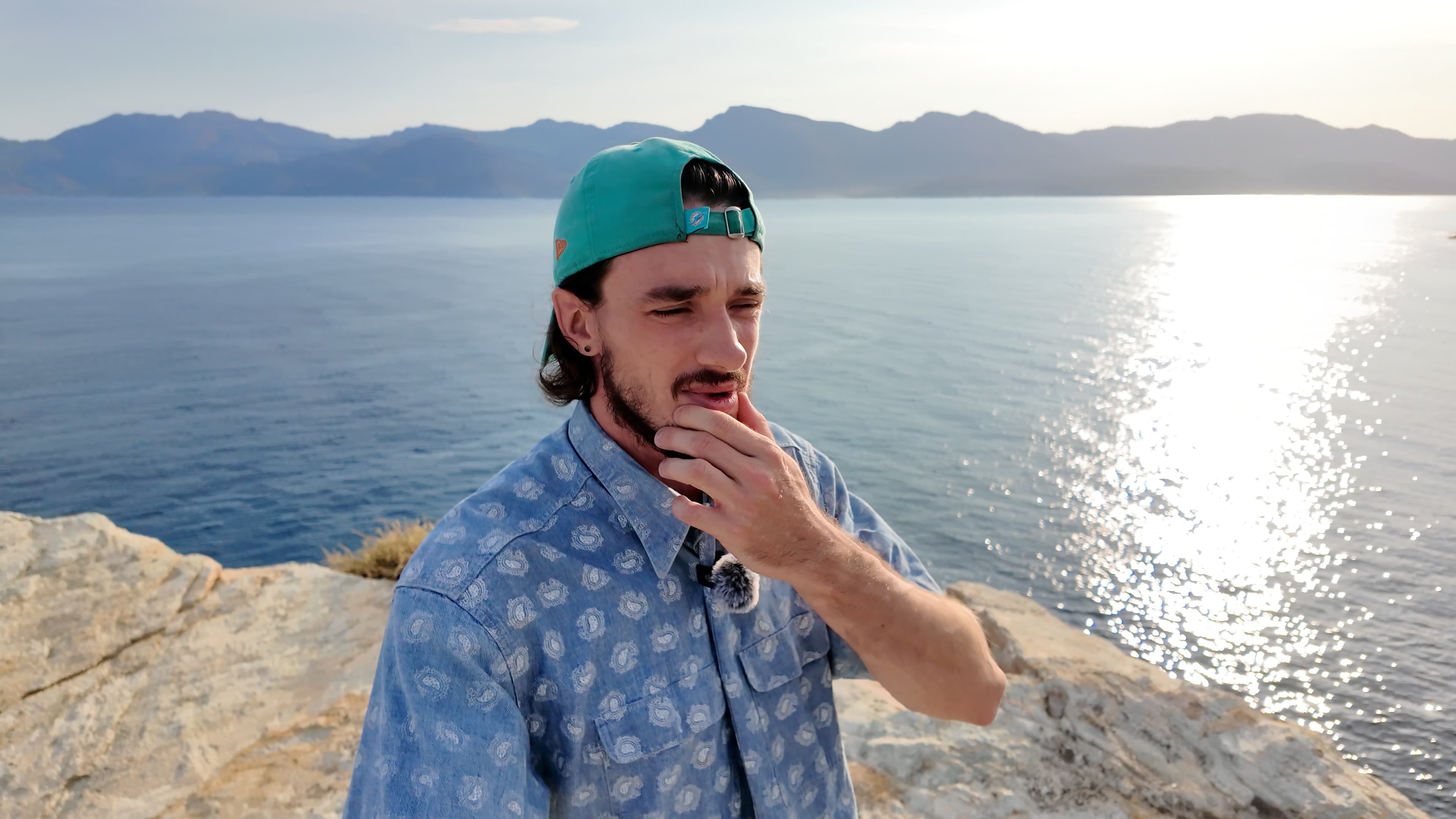 Ybelion (Samuel Le Parc) face à la mer Méditerranée, main devant la bouche dans un geste hésitant, montagnes en arrière-plan