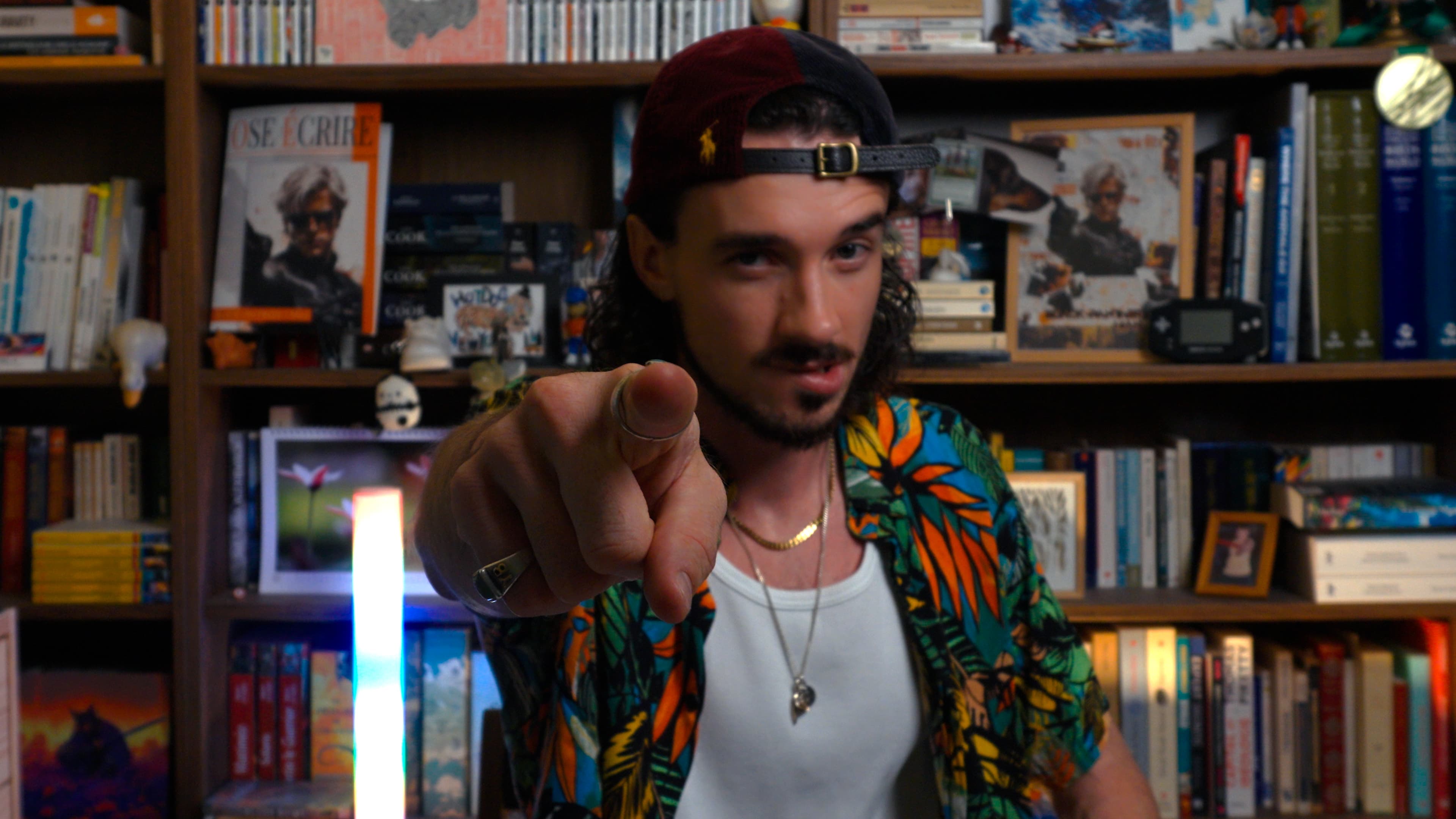 Ybelion (Samuel Le Parc), casquette à l'envers et chemise tropicale, pointant le doigt vers l'objectif devant sa bibliothèque