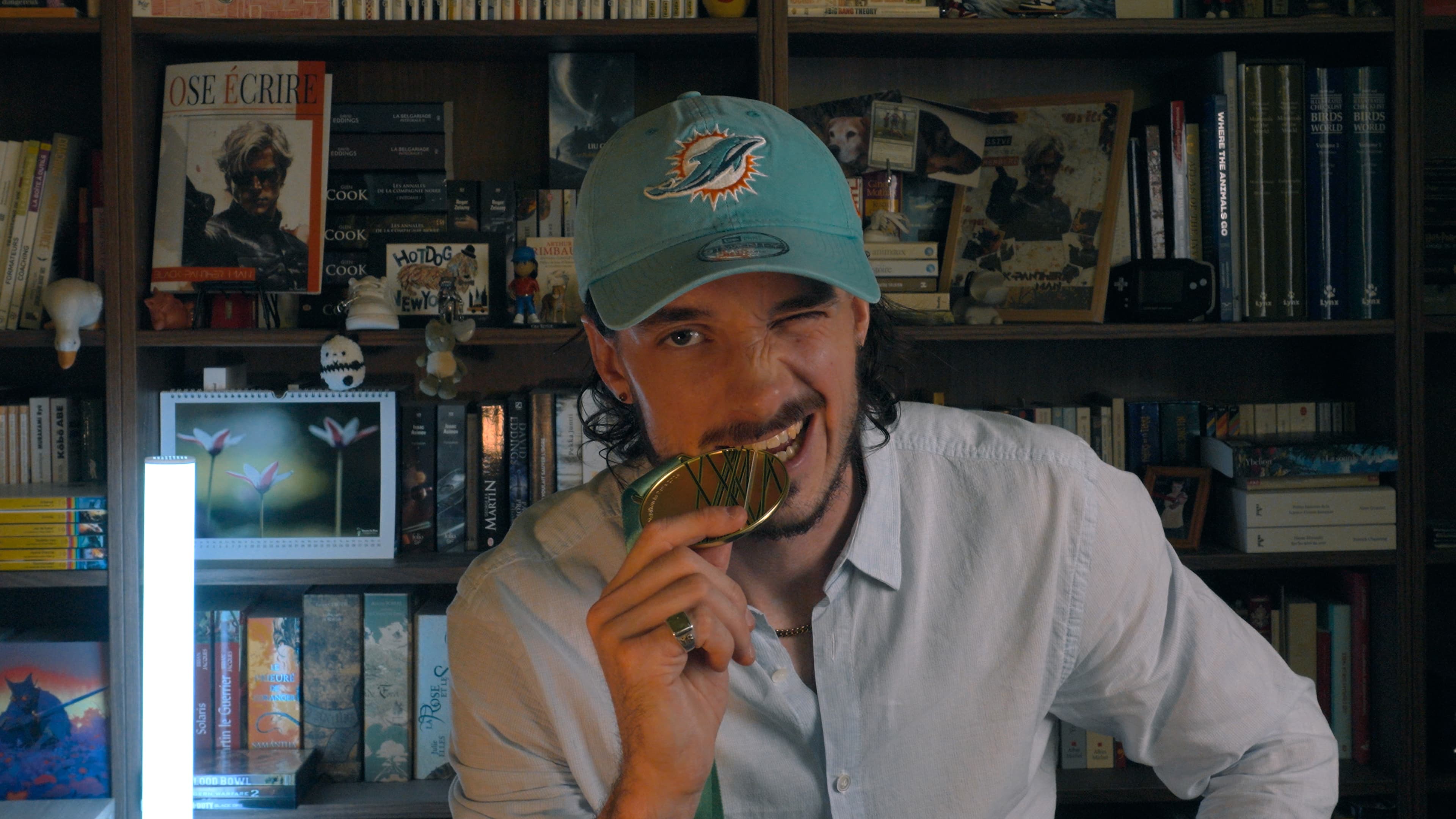 Ybelion (Samuel Le Parc), casquette turquoise des Miami Dolphins, mordant sa médaille dorée de marathon devant sa bibliothèque