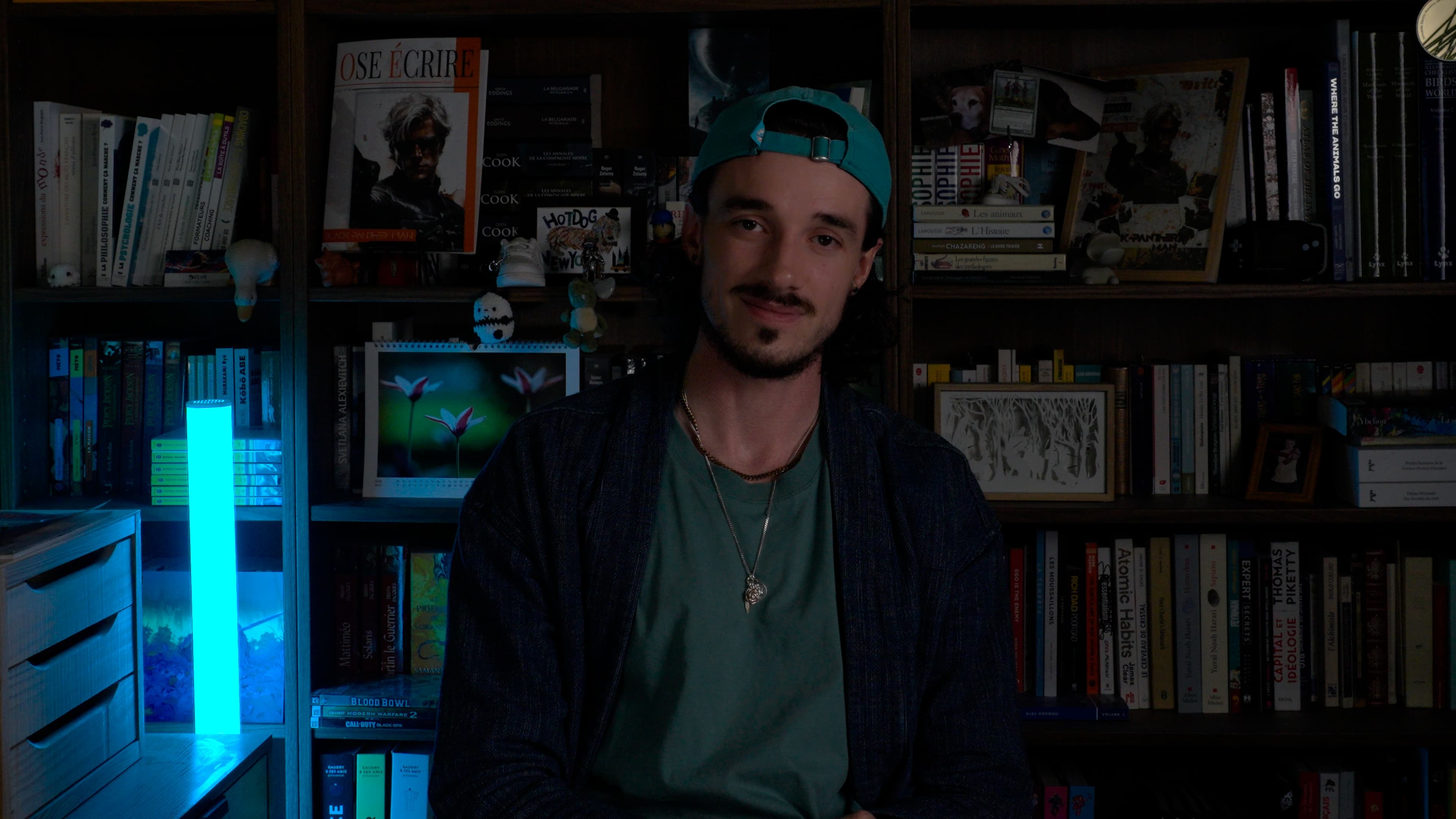 Ybelion (Samuel Le Parc), casquette à l'envers et t-shirt vert, assis dans son bureau tamisé devant ses étagères de livres