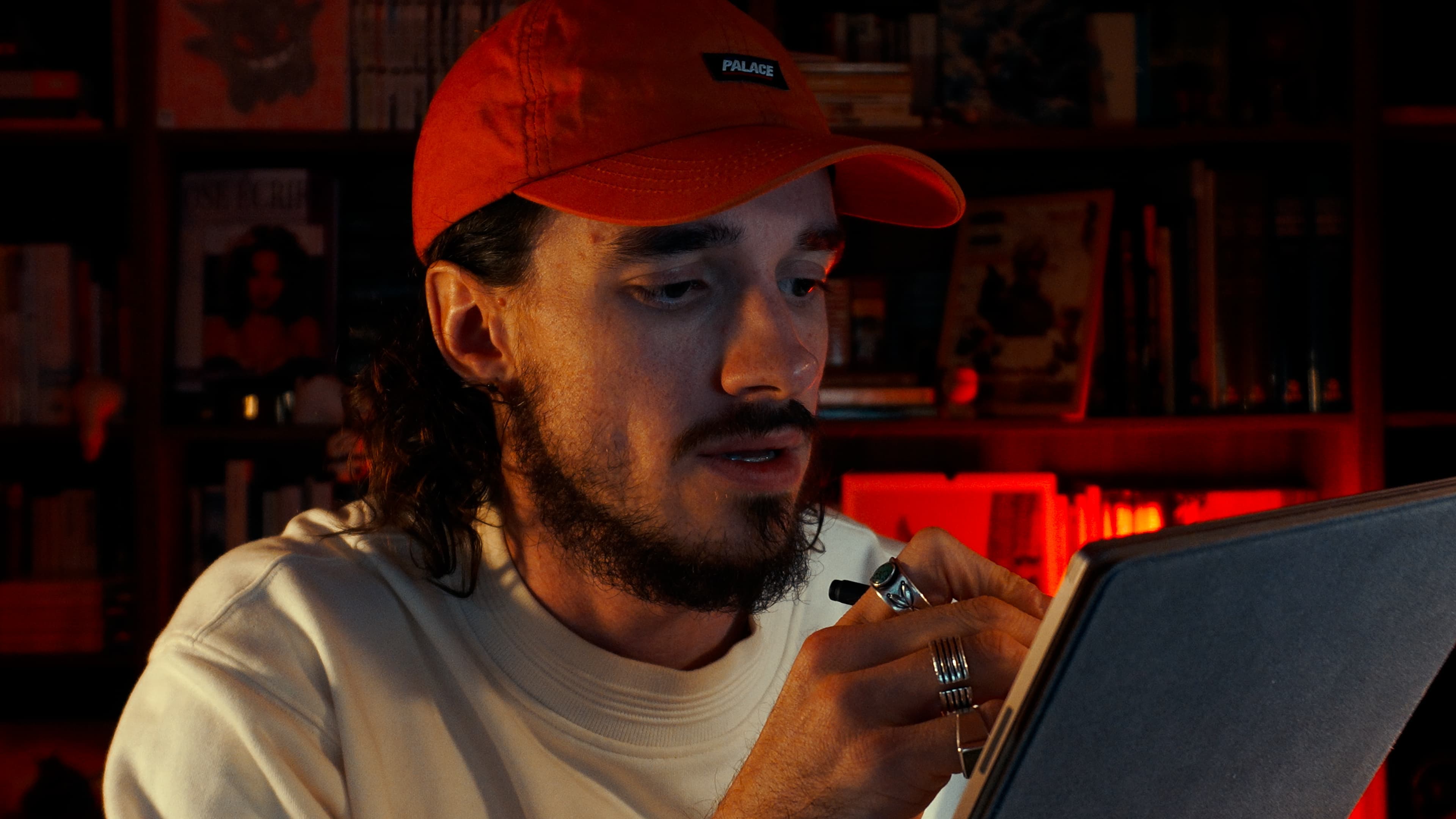 Ybelion (Samuel Le Parc), casquette rouge, écrit au stylet sur une tablette dans une bibliothèque éclairée en rouge.